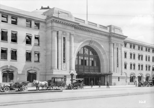 Une photographie en noir et blanc illustrant l’entrée d’un imposant édifice. Des automobiles et des carrioles tirées par des chevaux attendent en file, et des gens sont debout près de l’entrée. 