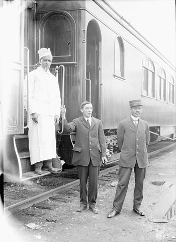 Photographie en noir et blanc de trois hommes près d’une voiture ferroviaire. Un chef cuisinier se tient sur les marches d’accès au train, un autre tient la main-courante, et le troisième, un porteur, est légèrement à l’écart, à côté du train.