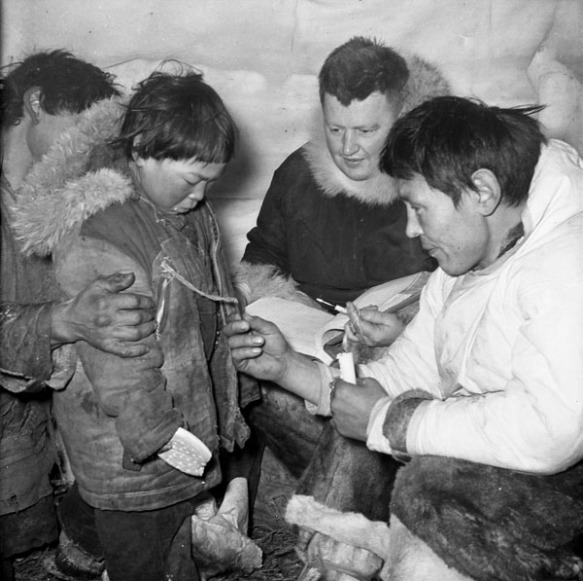 Une photographie en noir et blanc, prise dans un iglou, de deux hommes lisant un numéro de disque attaché au parka d’un jeune garçon.
