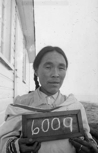 Une photographie en noir et blanc d’une femme inuite tenant un petit tableau sur lequel est écrit le numéro 6009.