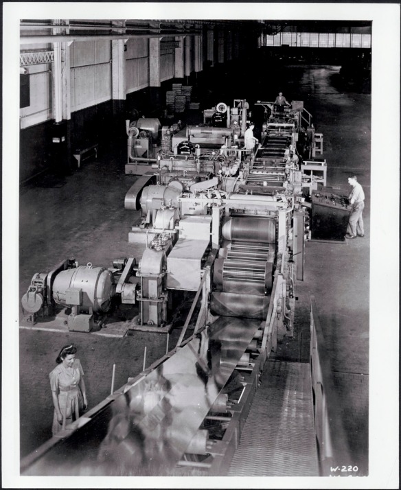 Photographie noir et blanc montrant des travailleurs manœuvrant une machine servant à rouler l’acier et à le transformer en multiples panneaux.