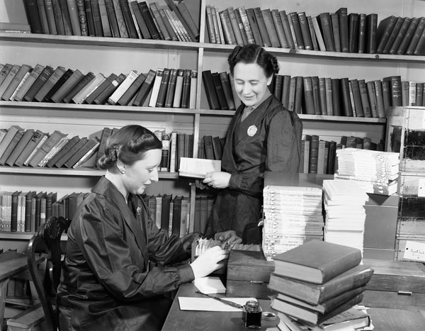 Photographie en noir et blanc de deux femmes dans une bibliothèque. L’une examine un catalogue sur fiches et l’autre tient un livre et regarde l’autre femme travailler.