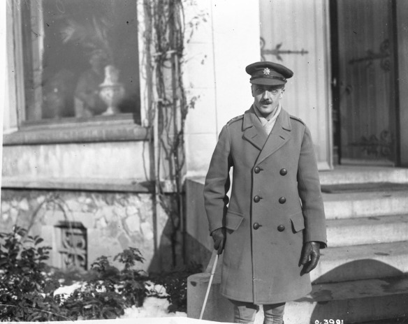 Photo noir et blanc d’un officier devant un bâtiment; il porte une casquette d’officier et tient un bâton de marche dans sa main droite.