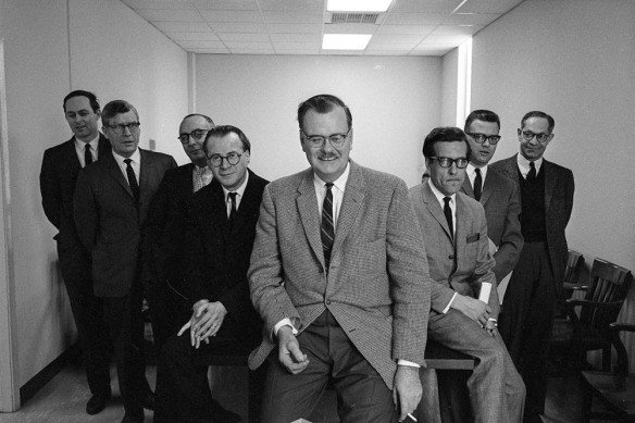 Photo noir et blanc de huit hommes en complets, souriant devant l’objectif, dans une salle de conférence aux murs clairs. 