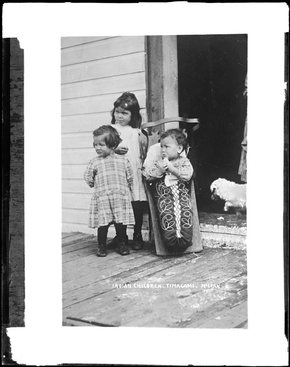 Photographie noir et blanc de trois enfants. Le plus jeune est installé dans un tikinagan au motif de broderie fleurie.