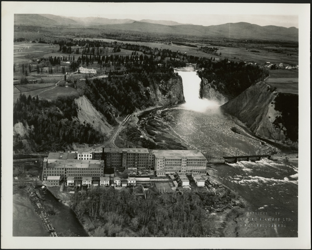 Une photographie aérienne noir et blanc montrant une usine près d’une rivière, avec une imposante chute à l’arrière-plan.