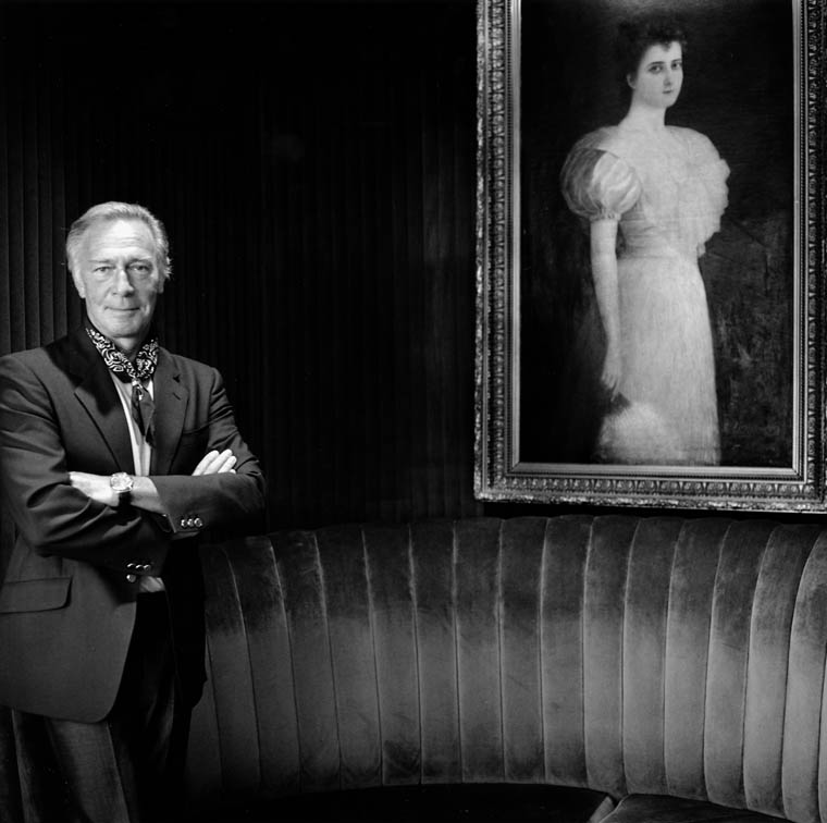 Photo noir et blanc de Christopher Plummer en complet, debout à gauche, avec les bras croisés. Une grande peinture encadrée, montrant une femme vêtue d’une robe et tenant un éventail, est accrochée sur la droite. 