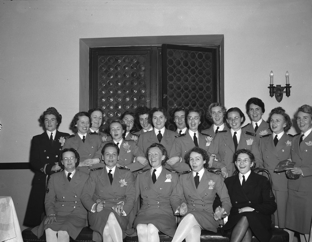 Photo noir et blanc d’un groupe de femmes en uniforme souriant à l’objectif. Deux des femmes portent un costume plus sombre. Celles au premier rang sont assises et se tiennent par la main. Les autres sont debout derrière; plusieurs se tiennent par le bras.