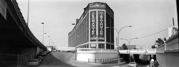 Photo noir et blanc d’un grand bâtiment d’usine à une intersection routière. Un viaduc reliant le bâtiment à une route se trouve du côté droit de l’image. Du côté gauche, on peut voir une file de voitures garées. 