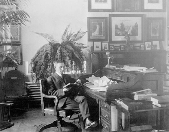 Photo noir et blanc d’un homme moustachu portant un costume sombre et des bottes. Il est assis sur une chaise de bois et lit un livre, près d’un bureau de bois recouvert de papiers. À l’arrière-plan, on voit de grandes plantes, un mur présentant de nombreuses images encadrées et un manteau de cheminée. Le fauteuil de campagne en cuir du général Wolfe est appuyé contre le mur, à la droite de l’homme.