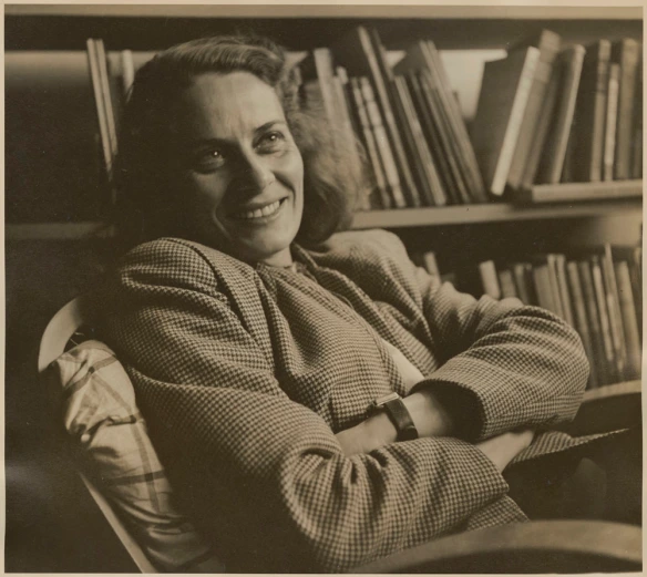 Photographie sépia montrant une femme blanche aux cheveux foncés. Elle est assise devant une étagère remplie de livres, ses bras sont croisés et un sourire traverse son visage.