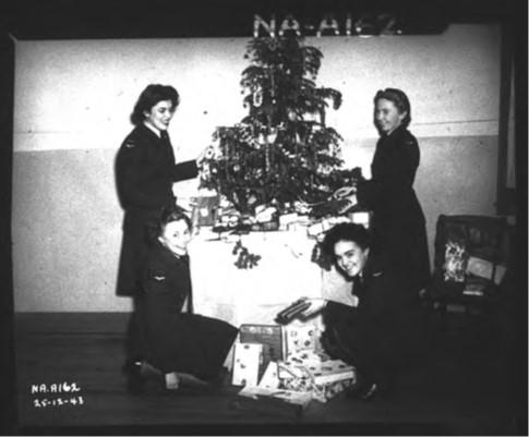 Quatre femmes en uniforme, debout ou agenouillées près d’un sapin de Noël posé sur une table. Des cadeaux emballés se trouvent sur le plancher et au pied de l’arbre.